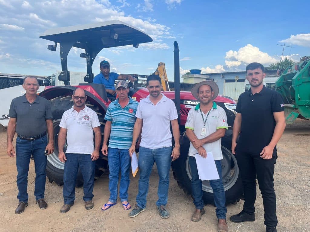 Sete pessoas reunidas em frente a um trator agrícola vermelho, durante a entrega do trator cedido pela Prefeitura de Córrego Fundo ao Conselho